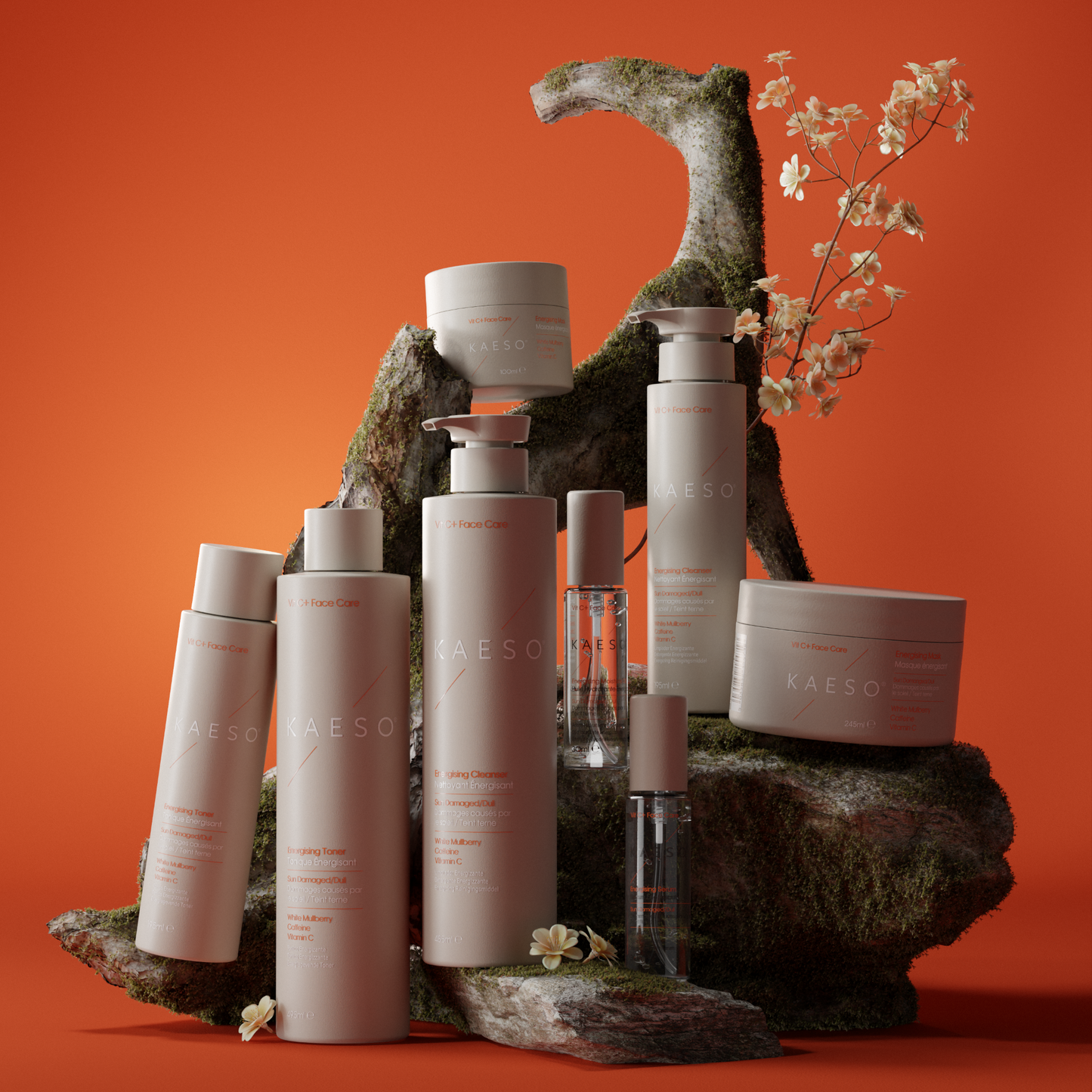 A collection of KAESO skincare bottles, jars, and a small travel sample vial arranged on mossy rocks against a solid orange background. The products are off-white with light pink text, and the scene is styled with small white flowers.