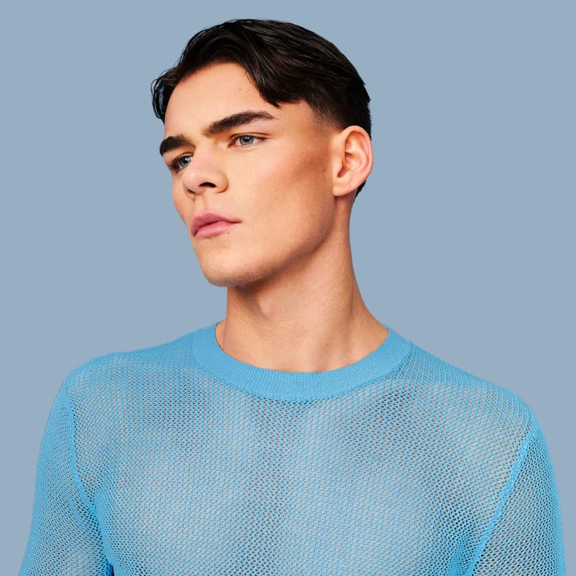 A close-up portrait of a young man wearing a light blue open-knit mesh crewneck sweater against a solid light blue-gray background.