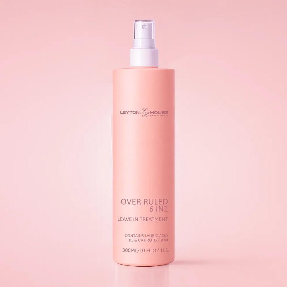 A 300ml bottle of Leyton & Holmes Over Ruled 6-in-1 Leave In Treatment spray, in a soft peach-pink color with a white spray nozzle, set against a matching solid pink background.