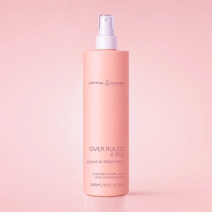 A 300ml bottle of Leyton & Holmes Over Ruled 6-in-1 Leave In Treatment spray, in a soft peach-pink color with a white spray nozzle, set against a matching solid pink background.