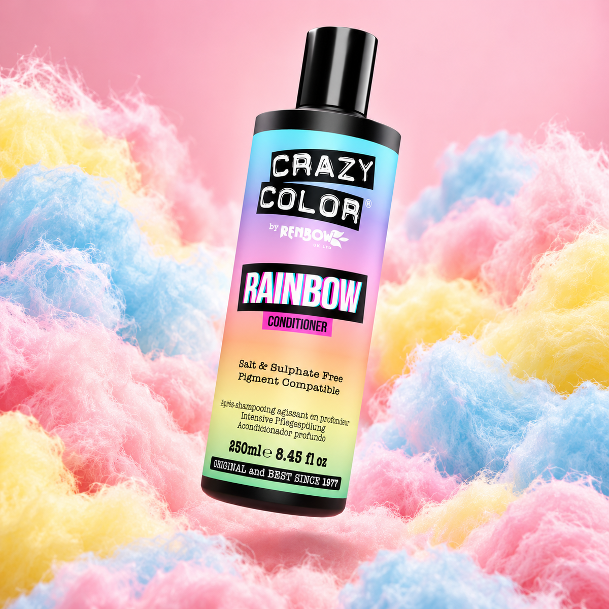 A bottle of Crazy Color by Renbow Rainbow Conditioner stands upright against a background of fluffy pastel cotton candy in pink, blue, and yellow. The bottle has a black cap and a vibrant rainbow gradient label with black and white text.