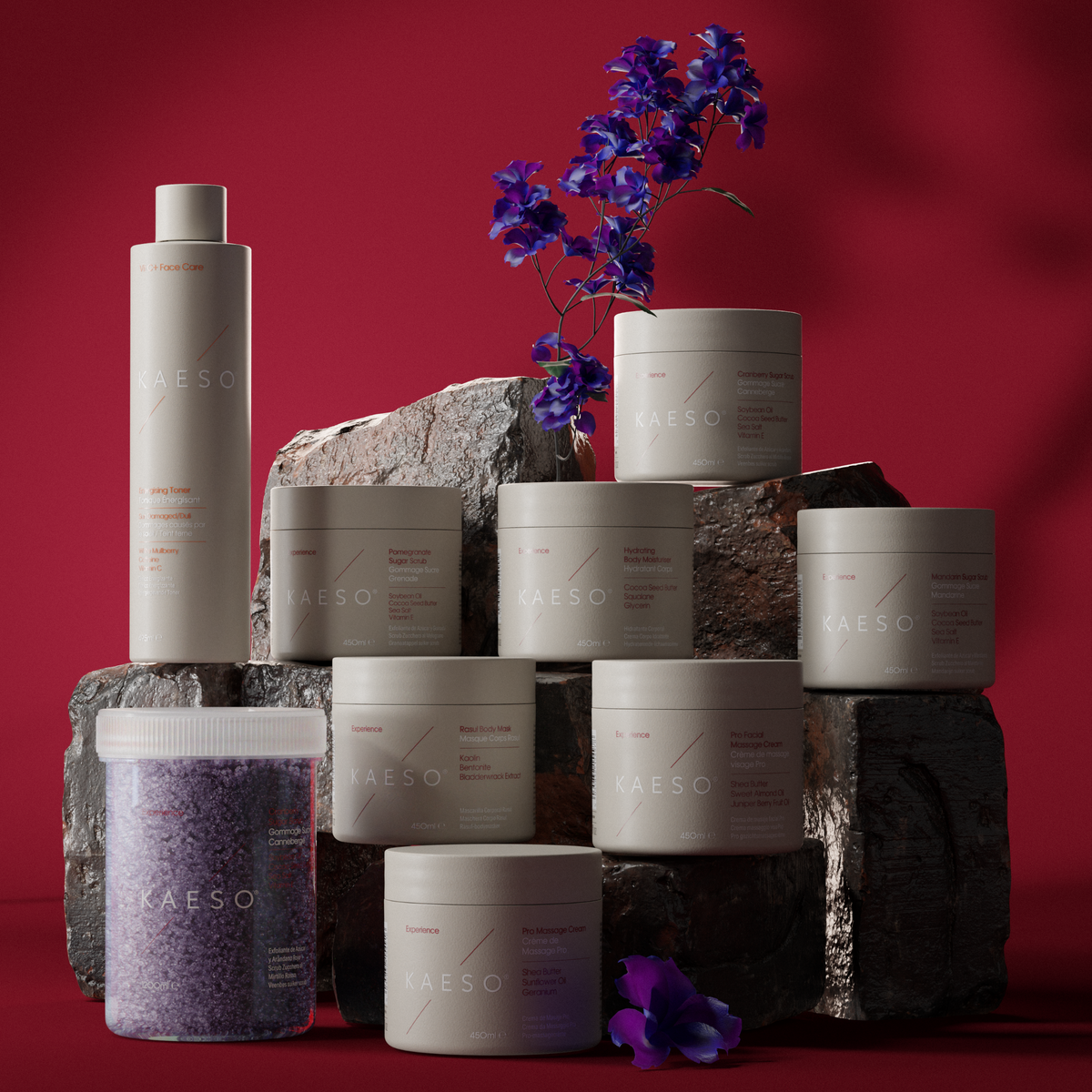 A collection of KASEO skincare products in matte off-white packaging, including a tall bottle and multiple jars, styled on dark textured stone blocks against a deep red background with purple flowers
