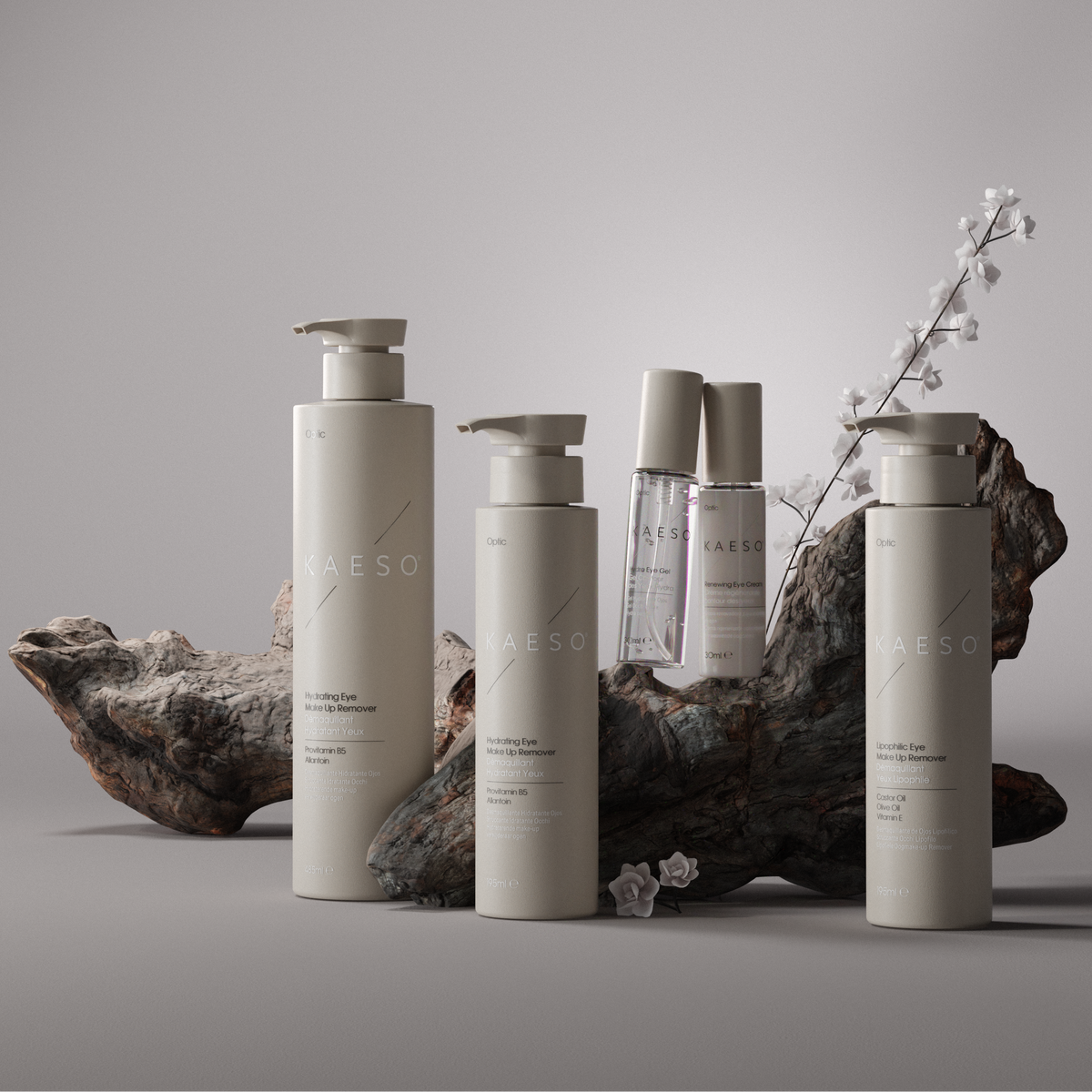 A set of six KAESO skincare and haircare bottles in a neutral matte beige and clear finish, arranged on a dark piece of driftwood against a solid gray background.
