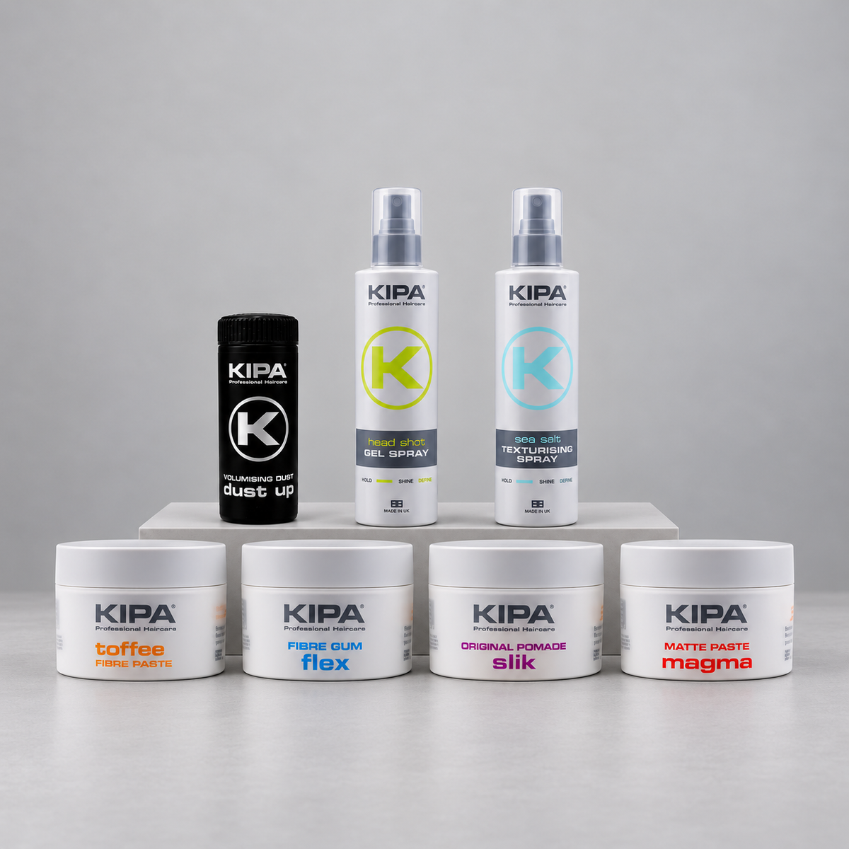 A set of KIPA hair styling products arranged on a light gray studio background, including a black dust-up container, two white textured spray bottles, and four white jars labeled coffee, fibre gum flex, original pomade silk, and matte paste magma.