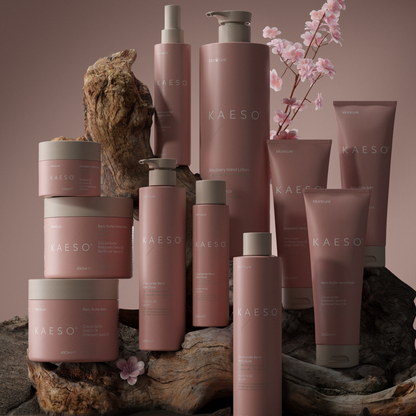 A collection of KAESO haircare products in coordinated dusty pink packaging, arranged on rustic driftwood and branches against a warm taupe-brown studio background. Visible products include bottles, jars, and tubes with pump and flip-top closures.