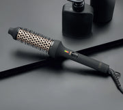 A black ionic heated brush with a ceramic barrel, laid on a flat surface, with a power cord attached.