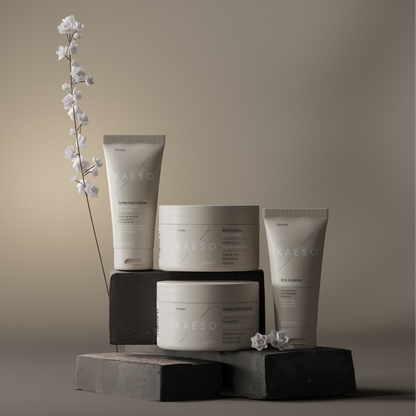 Four MAESO skincare containers arranged on dark geometric blocks against a warm taupe studio background, with a small flowering stem and flowers as styling props.