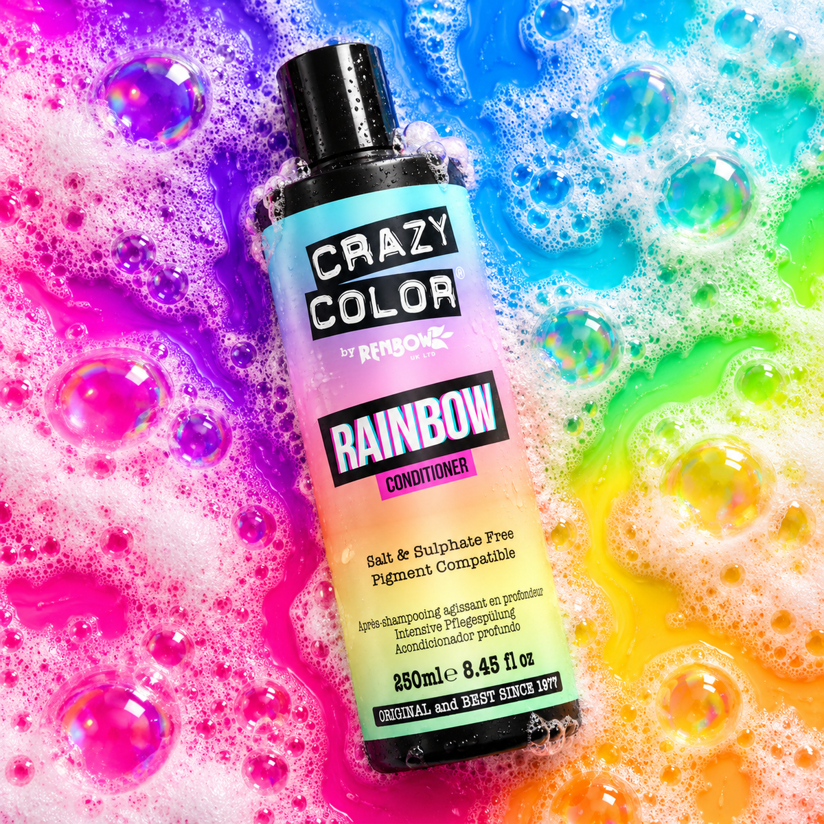 A bottle of Crazy Color by Renbow's Rainbow Conditioner lies diagonally on a bed of bright, multicolored soap bubbles and foam. The bottle features a black cap and a label that transitions from purple to pink to yellow to green, representing the 'Rainbow' color identity.